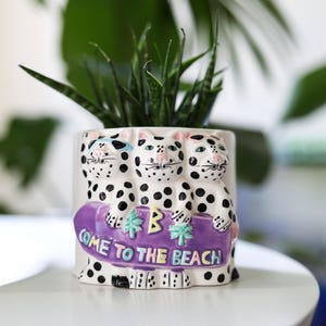 Könnte beinhalten: Weißer Keramik-Pflanzer in Form von drei Katzen mit Sonnenbrillen, die ein lilafarbenes Surfbrett mit dem Text "Come to the Beach" und Palmen halten.