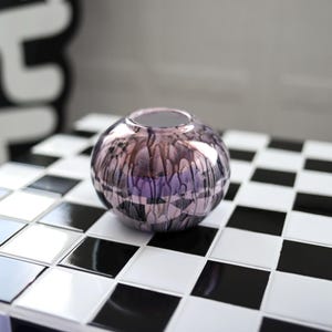 Könnte beinhalten: Eine kleine, runde, metallische Vase in Lila und Silber mit einer strukturierten Oberfläche steht auf einer schwarz-weißen Schachbrett-Fliesenfläche.