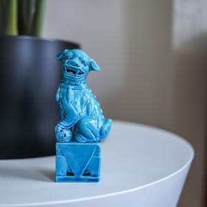 May include: A blue ceramic foo dog figurine sitting on a blue base. The foo dog is in a sitting position with its head turned to the side and its mouth open.