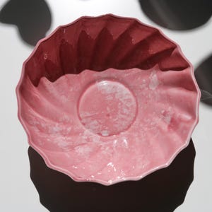 May include: A pink ceramic bowl with a scalloped edge and a circular indentation in the center. The bowl has a two-tone effect, with a darker shade of pink at the top and a lighter shade at the bottom. The bowl is likely used for serving food.