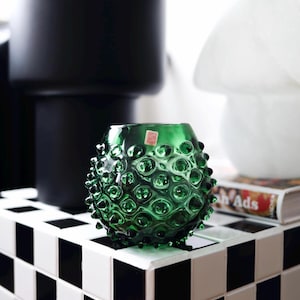Peut inclure: Un vase en verre vert avec une surface texturée en forme de bulles. Le vase est rond et repose sur une surface à carreaux noirs et blancs. Un pot noir et un objet décoratif blanc sont en arrière-plan.