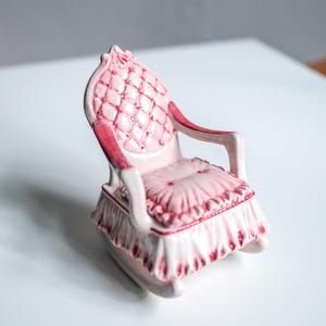 Könnte beinhalten: Kleiner, rosa Keramik-Schaukelstuhl mit gesteppter Rückenlehne und Sitzfläche. Der Stuhl hat einen gerüschten Rock und dunkelrosa Akzente. Das Design erinnert an ein Vintage-Puppenhaus oder einen Dekorationsartikel.