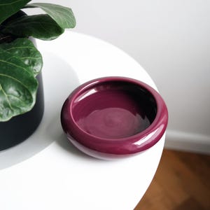 Könnte beinhalten: Eine tiefe, purpurfarbene Keramikschale mit abgerundetem Rand. Die Schale steht auf einem weißen Tisch.