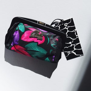 Könnte beinhalten: Eine schwarze Clutch mit floralem Muster, einem goldenen Verschluss und einem schwarz-weißen Anhänger. Der Anhänger hat einen schwarz-weißen Tierdruck.