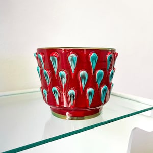 Peut inclure: Pot en céramique rouge avec un motif texturé de gouttes de couleur bleu sarcelle. Le pot est posé sur une étagère en verre.