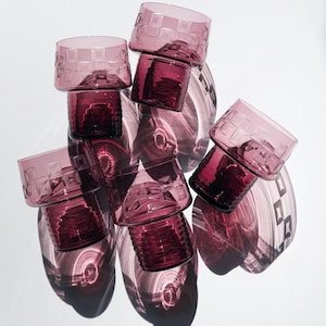Puede incluir: Colección de seis vasos de color amatista. Cada vaso tiene un patrón cuadrado en la parte superior y una base sólida. Los vasos están dispuestos en un grupo, proyectando sombras sobre una superficie blanca.