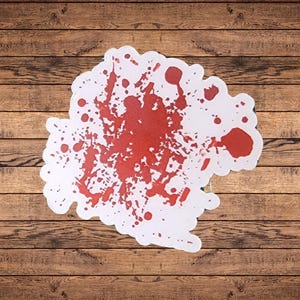 Puede incluir: Un recorte blanco de forma irregular con un diseño de salpicaduras de sangre roja. Las salpicaduras rojas varían en tamaño y densidad, creando un efecto visual llamativo. El recorte se coloca sobre un fondo de madera.