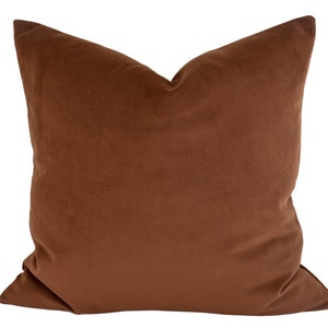 May include: A square, brown velvet pillow. The pillow has a soft, plush appearance and a solid color. The pillow is a warm, earthy tone, suitable for home decor. The pillow is a decorative item.