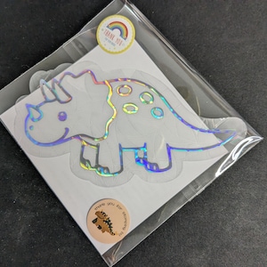 May include: A holographic triceratops sticker in a clear plastic bag. The sticker has a rainbow sheen and is outlined in a light colour. Two round stickers with text and a dinosaur graphic are also visible.
