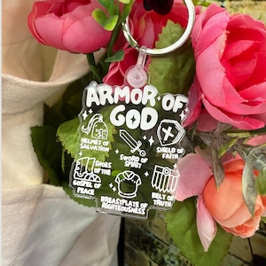 May include: Clear acrylic keychain with the words "Armor of God" and various religious symbols. The keychain includes a helmet, sword, shield, shoes, belt, and breastplate. The background features pink roses and greenery.