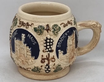 Puede incluir: Una jarra de cerveza de cerámica beige con un asa texturizada y un diseño detallado. La jarra presenta paneles azules con ilustraciones de castillos y las palabras "MAUS" y "DRACHEN-". Acentos de vid de uva verdes y marrones adornan la jarra.