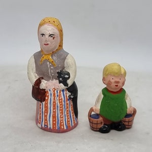 Könnte beinhalten: Zwei handbemalte Keramikfiguren, eine Frau in einem gelben Kopftuch und einem gestreiften Rock, die eine schwarze Katze hält, und ein Junge in einem grünen Hemd, der zwei Körbe hält.