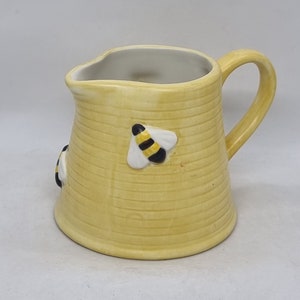 May include: A yellow ceramic creamer with a bee design. The creamer has a handle and a spout.