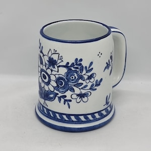 Puede incluir: Una taza de cerámica blanca con un diseño floral azul. La taza tiene un asa y una banda azul y blanca a rayas alrededor de la base.