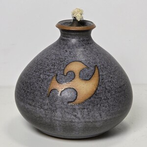 May include: A gray ceramic oil lamp with a brown and beige stylized design. The lamp has a round body and a small opening at the top for a wick.