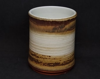 Puede incluir: Una taza de cerámica blanca con rayas marrones y naranjas. La taza tiene una forma ligeramente redondeada y un acabado liso y brillante.