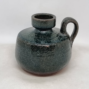 Può includere: Un vaso in ceramica verde scuro con manico e superficie testurizzata. Il vaso ha un corpo arrotondato e un'apertura leggermente svasata.