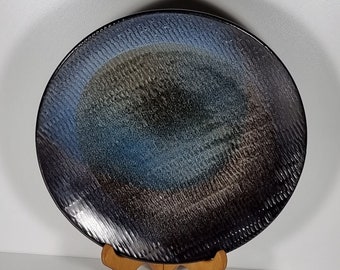 May include: A large, round ceramic plate with a dark blue, black and brown glaze. The plate has a textured surface and is displayed on a wooden stand.