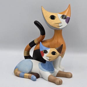 May include: A decorative figurine featuring two stylized cats. The larger cat stands upright with orange, black, and gold tones, while the smaller cat lies in front, colored in light blue, white, brown, and red, both with geometric facial features.
