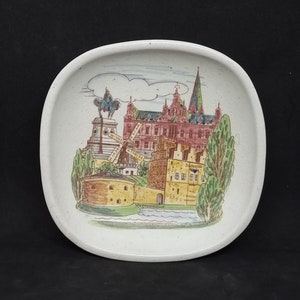 May include: A white ceramic plate with a hand-painted design of a castle with a red roof, green trees, and a blue river. The plate is oval-shaped and has a slightly raised rim.