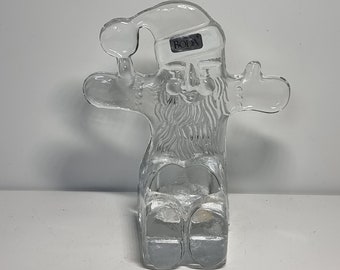 May include: Clear glass Santa Claus figurine with a beard and a hat. The figurine is sitting down with its arms outstretched. The figurine has a "BODA" label on its hat.