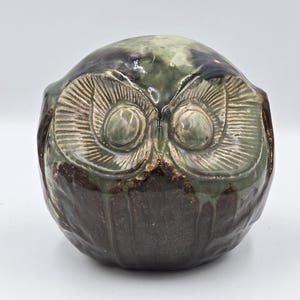 Może przedstawiać: Okrągła ceramiczna figurka sowy z błyszczącym szkliwem w odcieniach ciemnego brązu i przygaszonej szałwiowej zieleni. Sowa posiada duże, wyraźne okrągłe oczy ze szczegółowymi wygrawerowanymi liniami oraz gładki, zaokrąglony kształt korpusu.