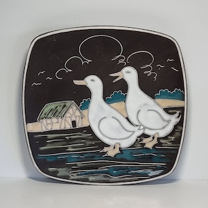 May include: A brown ceramic wall plate with a white farmhouse scene. Two white ducks are walking in a pond with a small brown house in the background. The plate has a dark brown background with white clouds and a white moon.