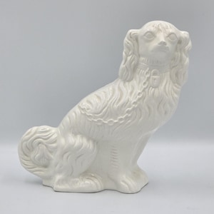Puede incluir: Figura de perro de cerámica blanca en posición sentada. El perro tiene pelo largo y fluido y un collar detallado. La figura es de color blanco liso y tiene un acabado brillante. La cola del perro está rizada y sus orejas son largas y caídas.