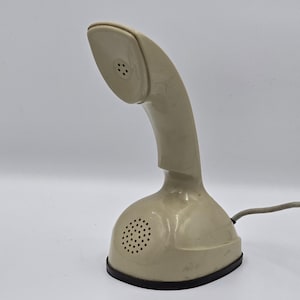 Peut inclure: Un combiné téléphonique beige monobloc avec une base au design incurvé et vertical. L'appareil présente une grille de haut-parleur sur le devant de la base et une grille assortie sur l'écouteur. Un cordon gris s'étend sur le côté de l'unité.