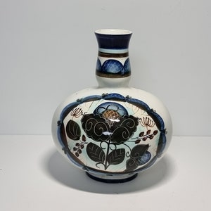 Puede incluir: Un jarrón de cerámica blanca con cuerpo redondo y cuello estrecho. El jarrón está decorado con un diseño floral azul y marrón.