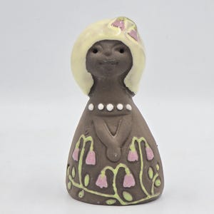 May include: A small ceramic figurine of a person with dark brown skin, a cream-coloured hairstyle with a pink bow, and a dark brown dress. The dress features a row of white dots at the neckline and a painted pattern of green vines with pink flowers.