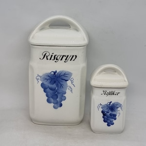 Op de afbeelding: Twee witte keramische potten met blauwe druivenmotieven en zwarte letters. De grotere pot heeft de tekst "Risgryn" en de kleinere pot heeft de tekst "Nejlikor".