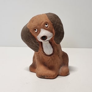 May include: A small ceramic dog figurine painted in brown and white. The dog is sitting with its head tilted to the side and its ears flopped down.