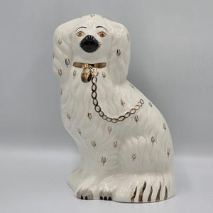 Könnte beinhalten: Eine weiße Keramik-Hundefigur mit glänzender Oberfläche und goldfarbenen Akzenten. Der Hund hat eine schwarze Nase, orangefarbene Augen und ein goldenes Halsband mit einer hängenden Kette. Kleine goldene Markierungen sind auf Körper und Ohren verteilt.