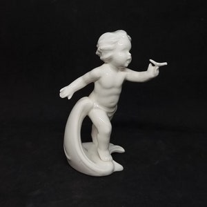 May include: A white porcelain figurine of a cherub holding a butterfly. The cherub is standing on a wave-like base.