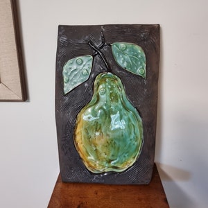 Puede incluir: Una placa de pared de cerámica con un diseño en relieve de una pera verde con hojas marrones. La pera tiene un esmalte amarillo y verde.