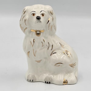 Puede incluir: Una figura de perro de cerámica blanca en posición sentada, con ojos marrones pintados, nariz negra y detalles dorados en las orejas, el pelaje y el collar. El perro tiene un acabado brillante y se representa con orejas largas y texturizadas.