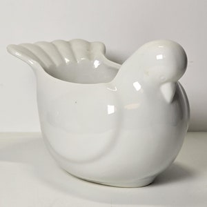 May include: A white ceramic dove-shaped planter. The planter has a smooth, glossy finish and a slightly rounded shape. It is ideal for displaying small plants or flowers.