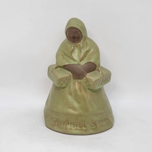 Può includere: Una statuetta in ceramica verde di una donna seduta con cesti su entrambi i lati. La statuetta è firmata "KRINGEL STAVA" sulla base.