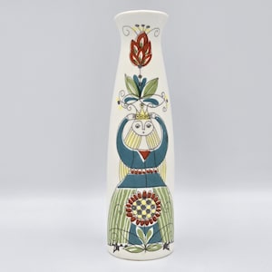 May include: A tall, off-white ceramic vase with a hand-painted design. The vase features a stylized figure with a blue top, green skirt, and yellow hair. Floral and geometric patterns in red, green, and yellow adorn the vase.