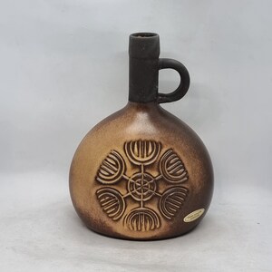 May include: A brown ceramic vase with a dark brown handle and a geometric design. The vase has a textured surface and a label that reads "Handgefertigt Keramik".