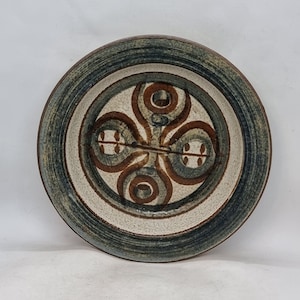 May include: A white ceramic plate with a blue and brown geometric design. The plate has a raised rim and a smooth surface.