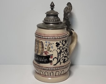 May include: A ceramic beer stein with a pewter lid and handle. The stein is decorated with a floral design and a barrel with the text "Glück Blume".