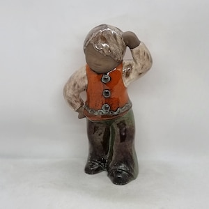 Puede incluir: Una figurita de cerámica de un niño con cabello castaño, que lleva una camisa blanca, un chaleco naranja con cuatro botones y pantalones marrones. El niño se está sosteniendo la mano en la cabeza, como si estuviera pensando.