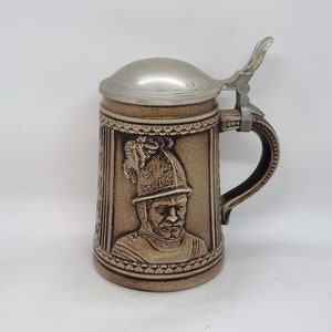 Op de afbeelding: Een bruine keramische bierkan met een tinnen deksel en een reliëfdesign van een man in harnas. De kan heeft een handvat en een gedetailleerd ontwerp rond de rand.