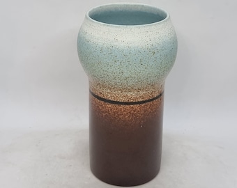 Könnte beinhalten: Eine Keramikvase mit einer hellblauen und weißen, gesprenkelten Glasur auf der oberen Hälfte und einer braunen Glasur auf der unteren Hälfte. Die Vase hat eine abgerundete Oberseite und einen zylindrischen Boden.