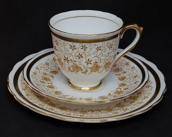May include: A white teacup and saucer set with a gold trim and a delicate floral pattern. The cup and saucer are both decorated with a repeating pattern of gold leaves and vines.
