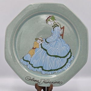 Puede incluir: Un plato decorativo octogonal verde claro con una ilustración azul y blanca de una mujer y un niño. La mujer está sentada, vestida con un vestido a rayas. El plato está inscrito con las palabras "Anna Karenina".