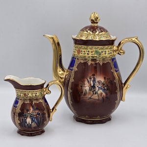 Pode incluir: Uma cafeteira e uma leiteira a condizer em porcelana com vidrado castanho escuro, acabamentos dourados e pegas ornamentadas. Ambas as peças apresentam cenas históricas detalhadas de figuras com uniformes militares montadas a cavalo.
