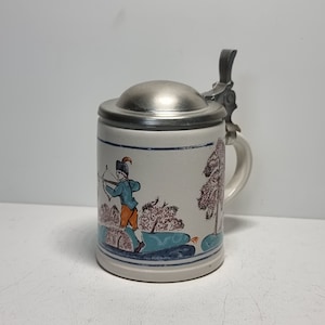 May include: A white ceramic beer stein with a pewter lid and handle. The stein features a hand-painted scene of a man in a blue and orange outfit with a bow and arrow, standing in a woodland setting.